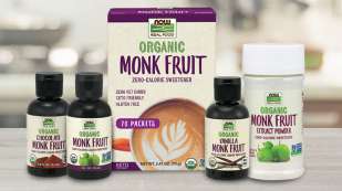 five Monk Fruit products on light colored counter - three small bottles of flavored liquid, one box of packets, one bottle of powdered Monk Fruit - all with light tan label with purple and green accents and lettering